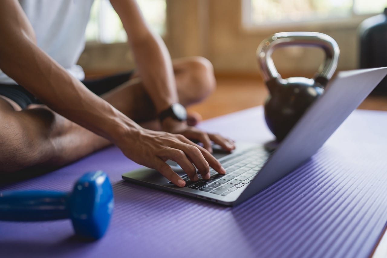 Young sportsman sitting on purple yoga mat and doing exercise sports workout online and looking video streaming on laptop indoor at the fitness gym. Online fitness training from gym concept. Be Strong Gym